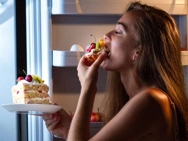 Hungry woman in pajamas eating sweet cakes at night near refrigerator.